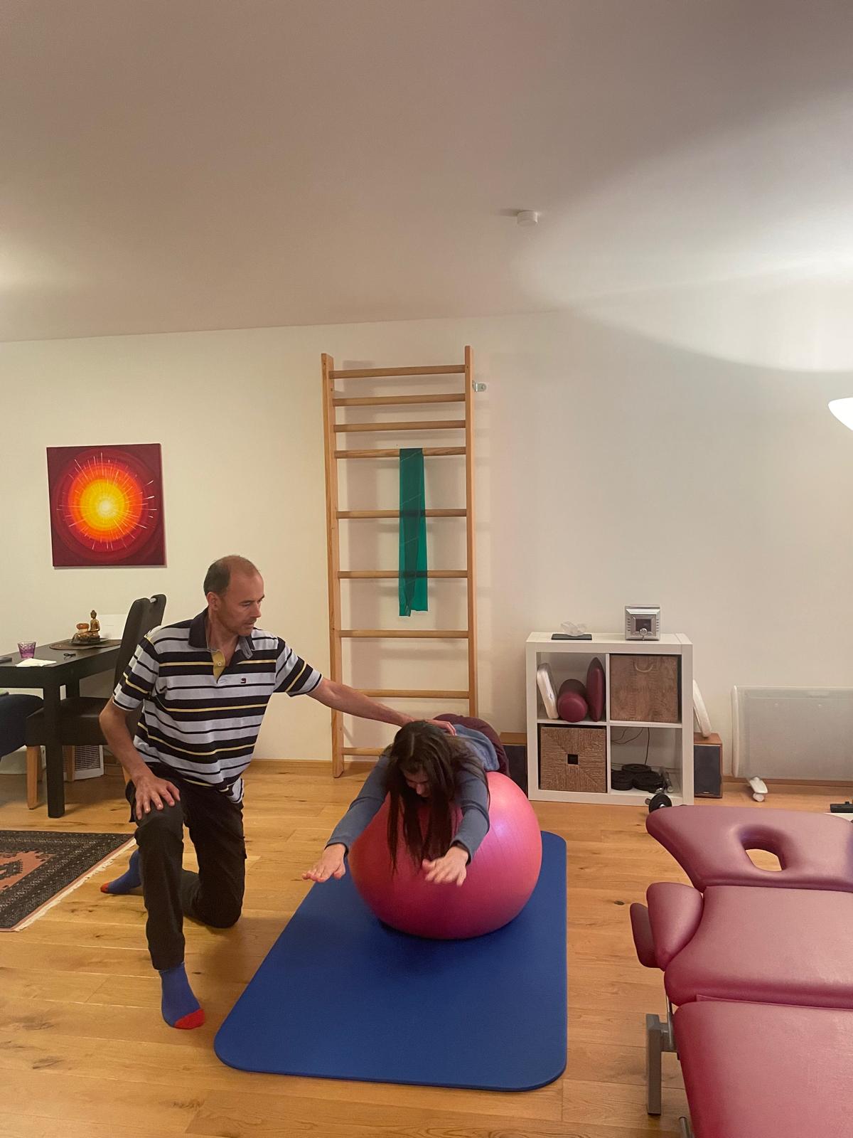 Eine Frau macht eine Übung auf einen Gymnastikball, Roland kniet daneben und kontrolliert die korrekte Ausführung der Übung. Im Hintergrund ist der Behandlungsraum mit diversen Geräten zu sehen.
