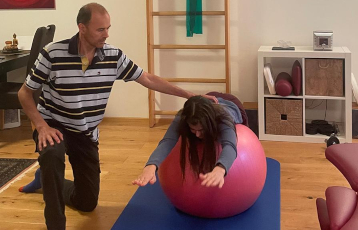 Eine Patientin macht eine Übung auf dem Gymnastikball, dabei steckt sie Ihre Arme nach Vorn. Roland kontrolliert ihre Haltung.