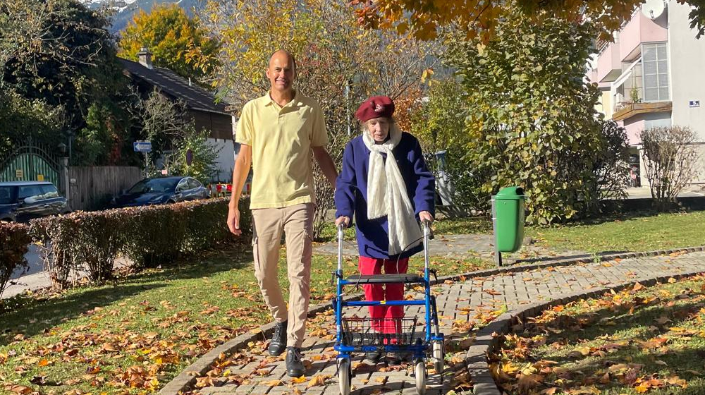 Roland und eine Dame gehen im Garten auf einen gepflasterten Weg. Es liegt herbstlich verfärbtes Laub auf der Wiese.