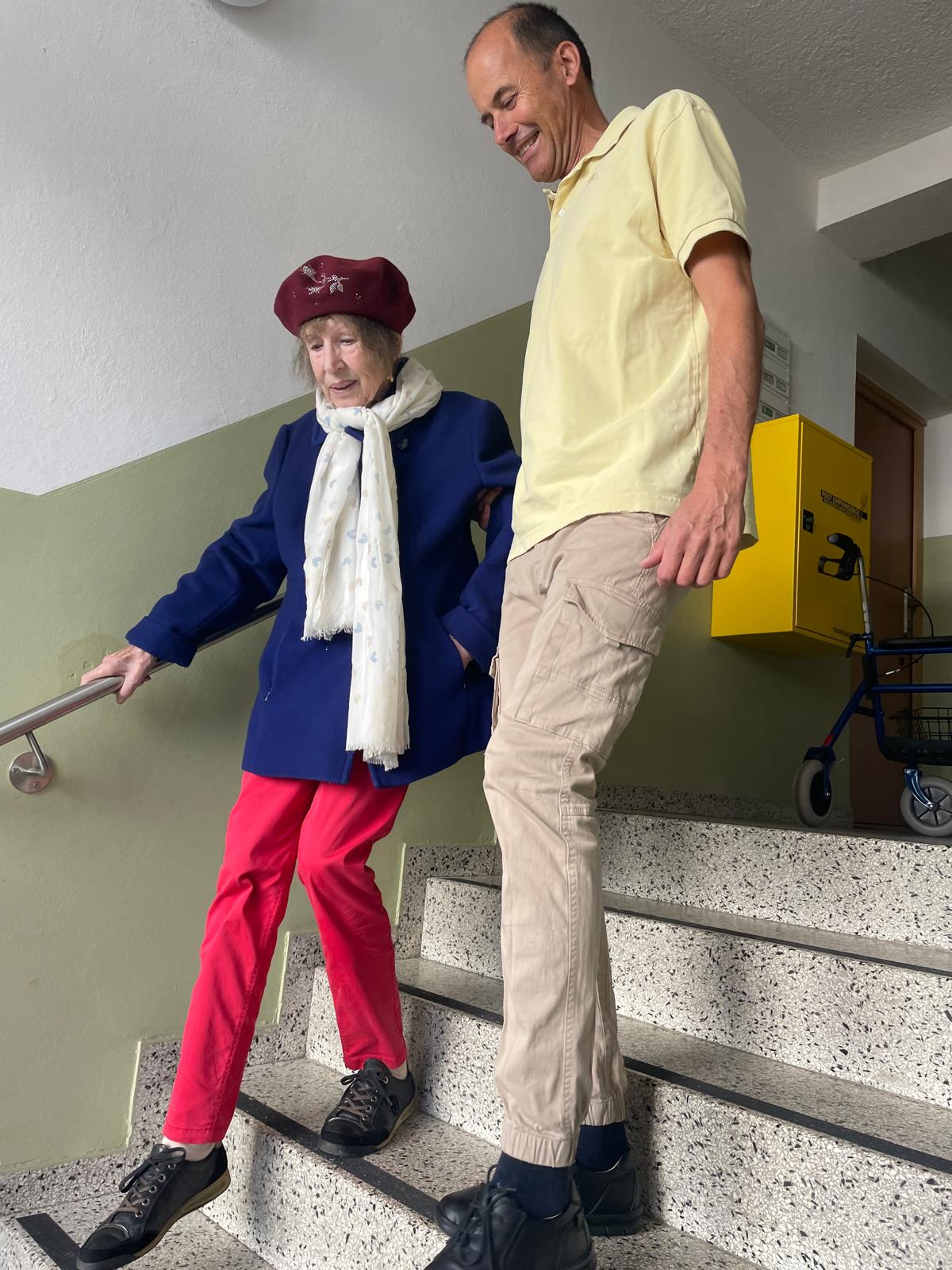 Roland und eine Dame gehen Treppen in einem Mehrfamilienhaus hinuter. Die Dame hät sich mit der rechten Hand am Treppengeländer fest, während sie zu ihrer linken bei Roland eingehakt ist. Beide haben ihren Blick nach unten gerichtet, um die Treppen anzusehen. Im Hintergrund am Podest der Treppen ist ein Rollator.